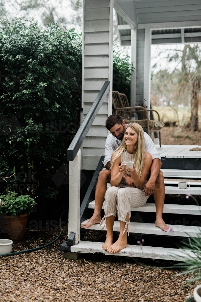 Man holding woman from behind as they sit on the steps of their porch - Australian Stock Image