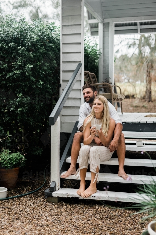 Man holding woman from behind as they sit on the steps of their porch - Australian Stock Image