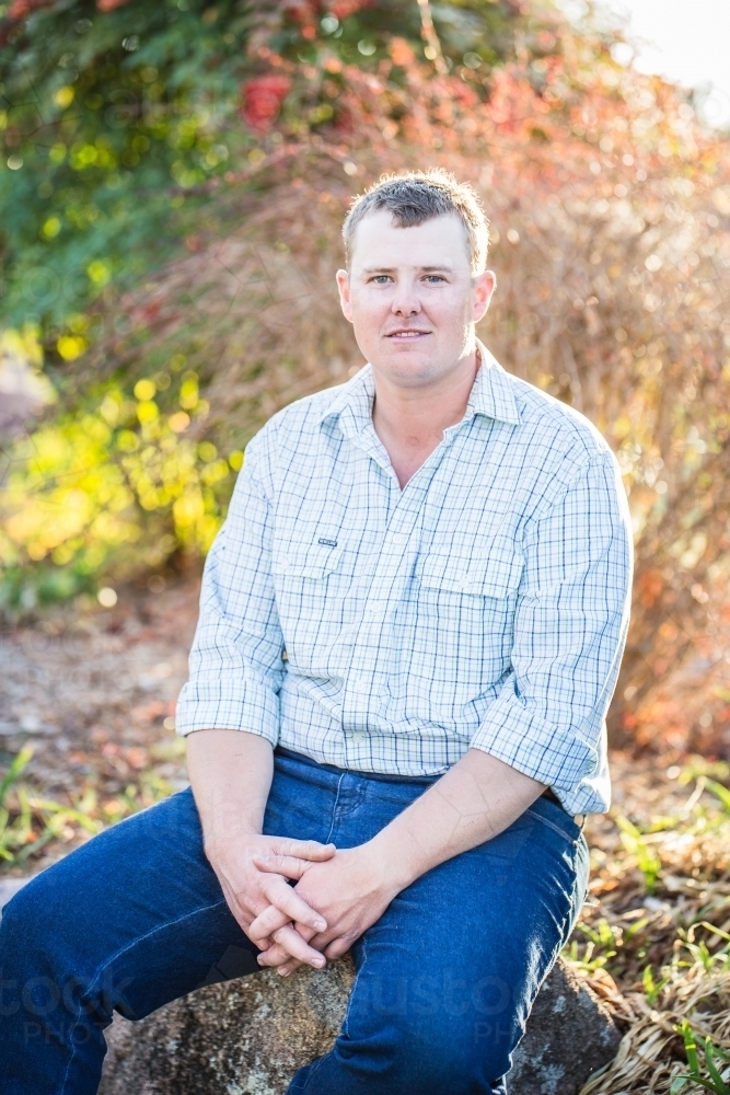 Man in 20s sitting on rock in garden with hands folded smiling - Australian Stock Image