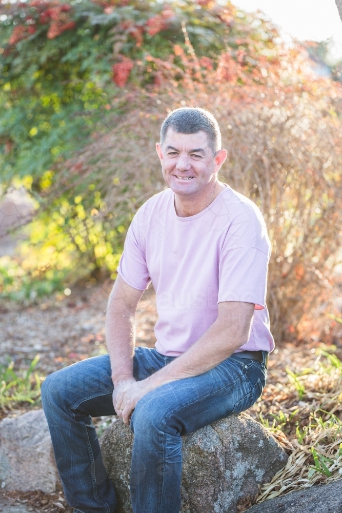 Man in 50s sitting on rock in garden smiling with dimples - Australian Stock Image