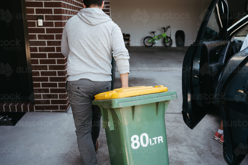 Man in a grey hoodie pushing a yellow-lidded wheelie bin - Australian Stock Image