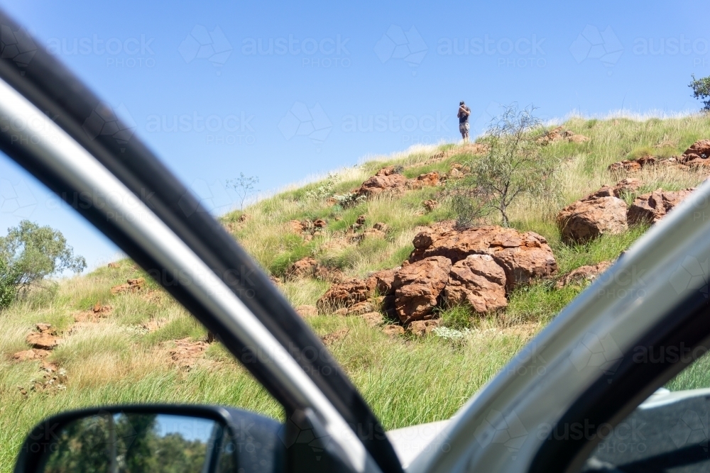 Man in distance standing on top of bushy rocky hill searching for service on his phone - Australian Stock Image