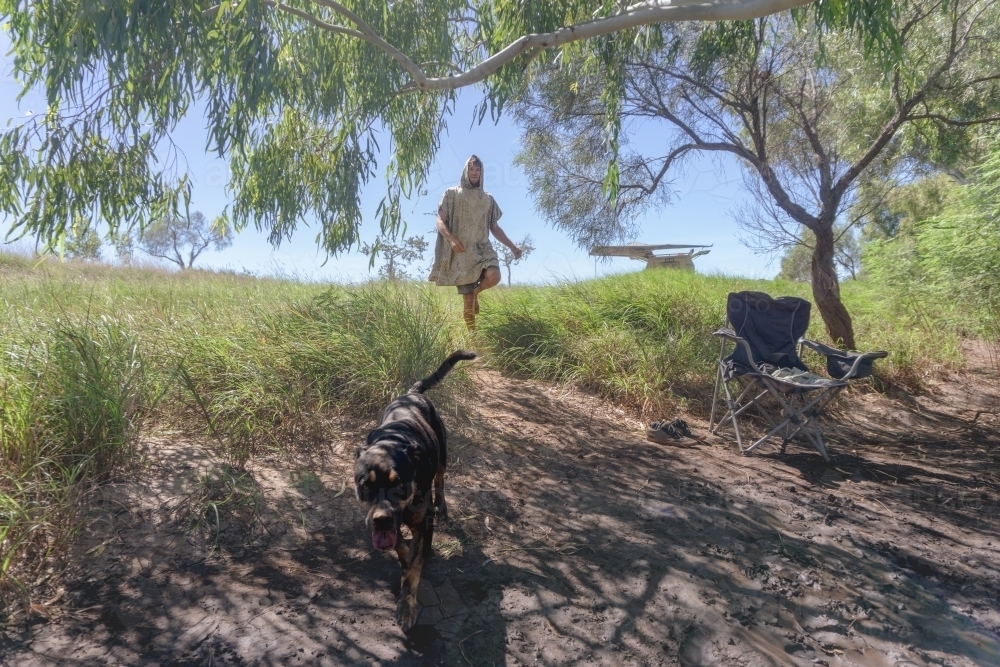 Man in hooded poncho towel walking down to riverside with dog leading the way, rugged bush of WA - Australian Stock Image