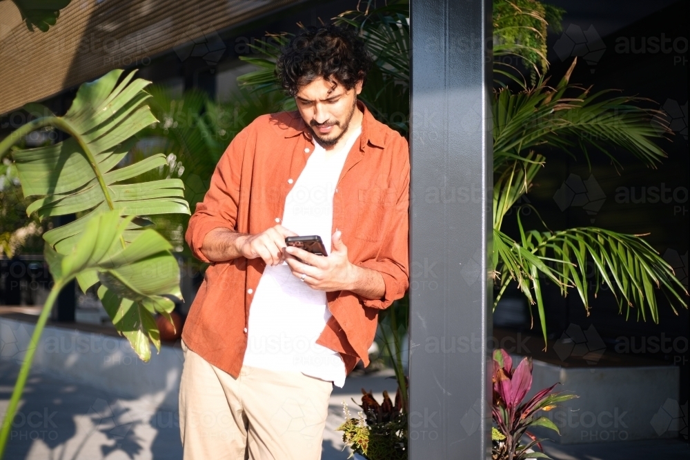 Man leaning against pole on phone with plants in the background - Australian Stock Image