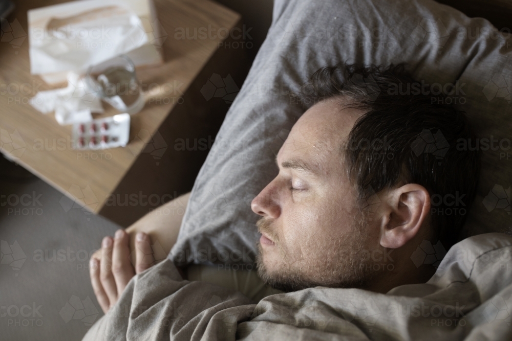 Man lying sick in bed with tissue box, medicine and glass of water - Australian Stock Image