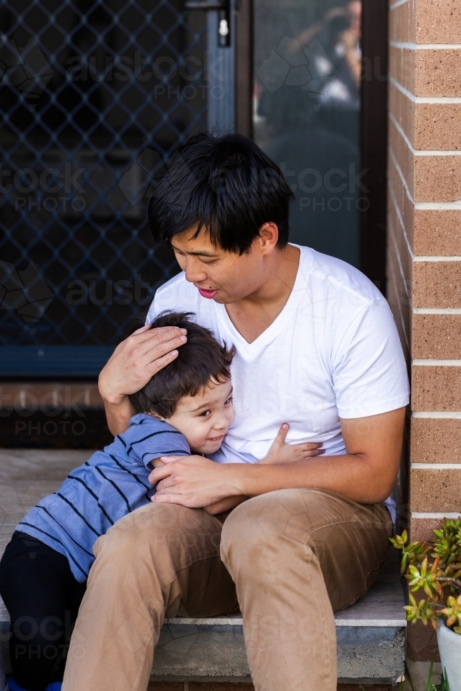 Man of Chinese ethnicity hugging mixed race son sitting on front steps of home - Australian Stock Image