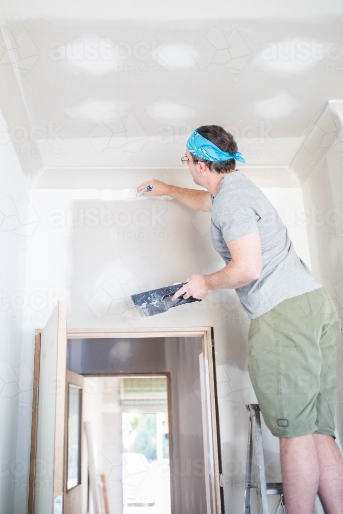 Man on ladder painting blank wall with white paint holding paint brush and tray - Australian Stock Image