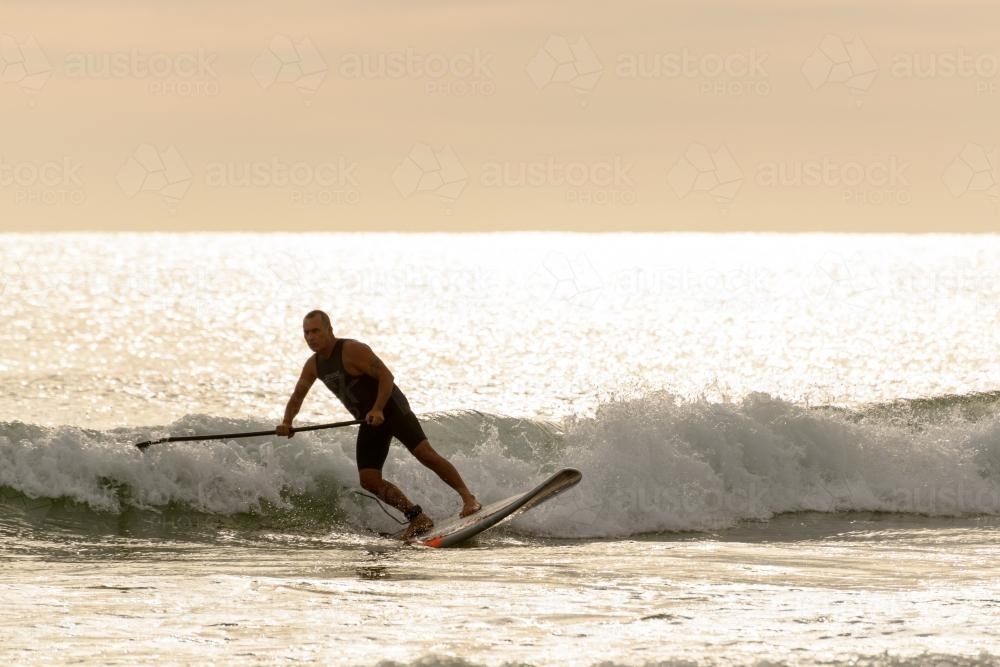 Man on paddleboard riding wave and backlit by dawn light on glistening water - Australian Stock Image