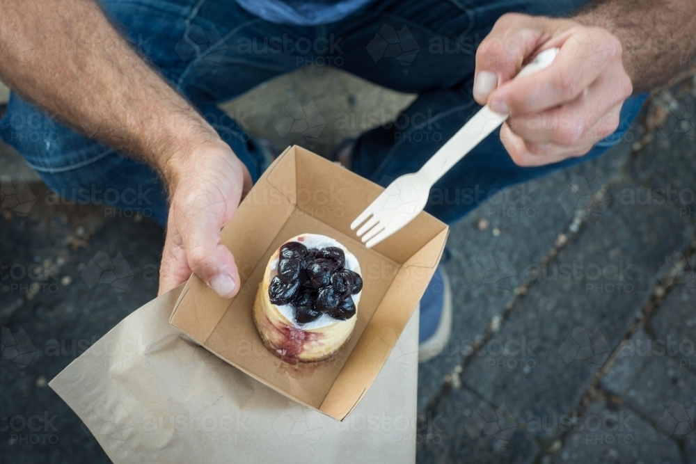 Man sitting on street curb eating bakery sweet - Australian Stock Image