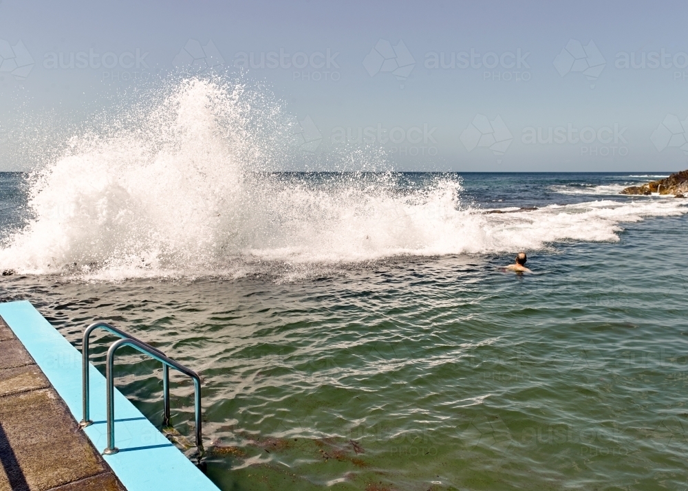 Man swimming in ocean pool with waves crashing over wall - Australian Stock Image