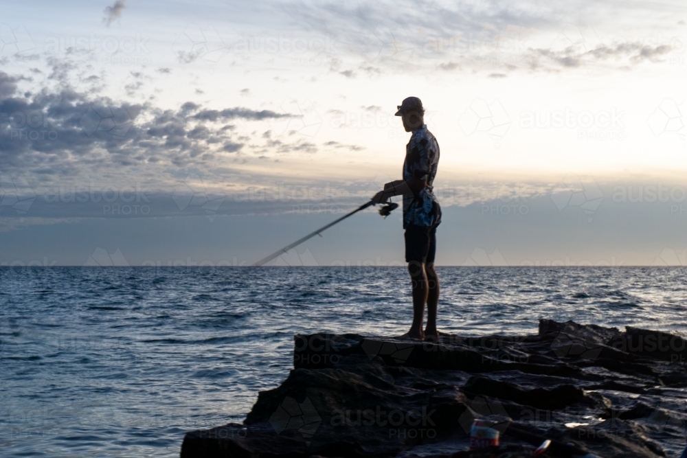 Image of Man wearing floral shirt fishing from rocks with sunset sky in the  background. Silhouette. - Austockphoto