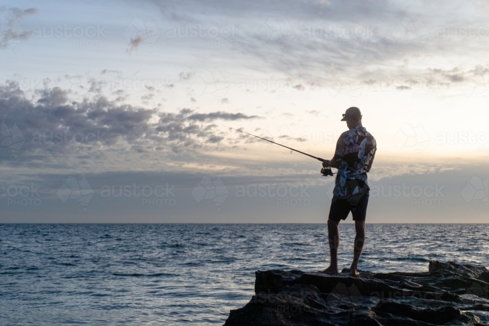 Man wearing floral shirt fishing from rocks with sunset sky in the background. Silhouette. - Australian Stock Image