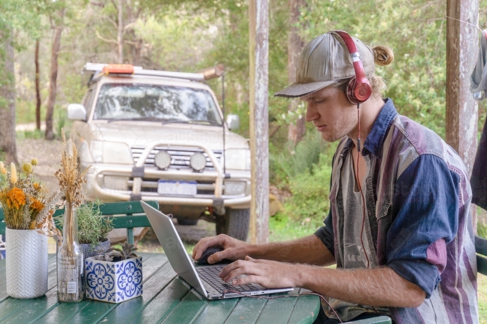 Man working remote on laptop with headphones on in the bush with trees and 4x4 car in background - Australian Stock Image