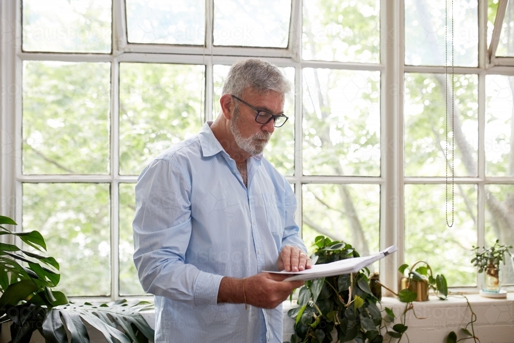 Mature businessman standing, reading near a paned window in open plan office - Australian Stock Image