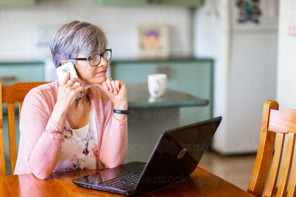 Mature woman on laptop at home using mobile phone to call for tech support - Australian Stock Image