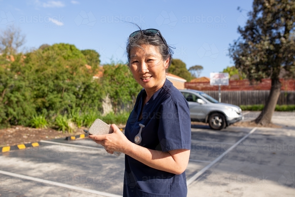 Medical nurse walking through a car park holding her phone smiling - Australian Stock Image