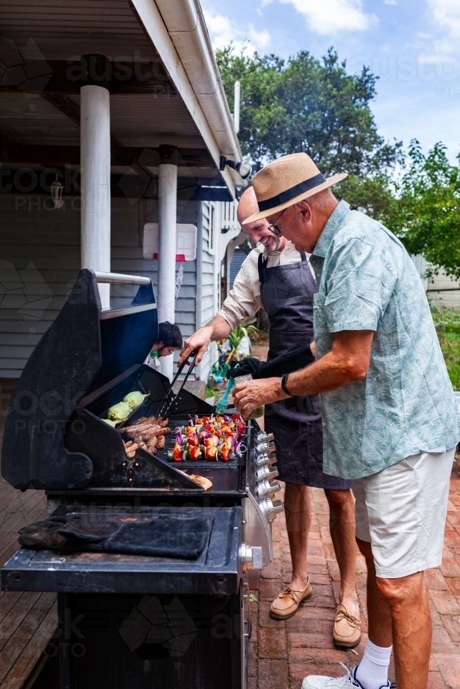 men cooking barbeque lunch in backyard together for midday meal with family - Australian Stock Image