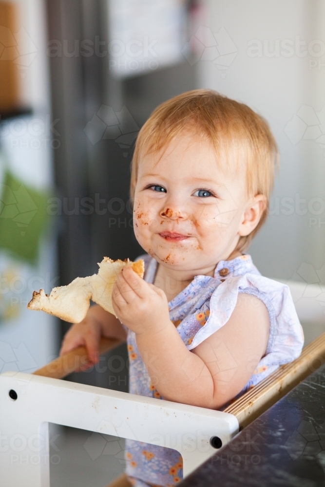 Messy baby girl eating vegemite toast in kitchen with cheeky smile - Australian Stock Image