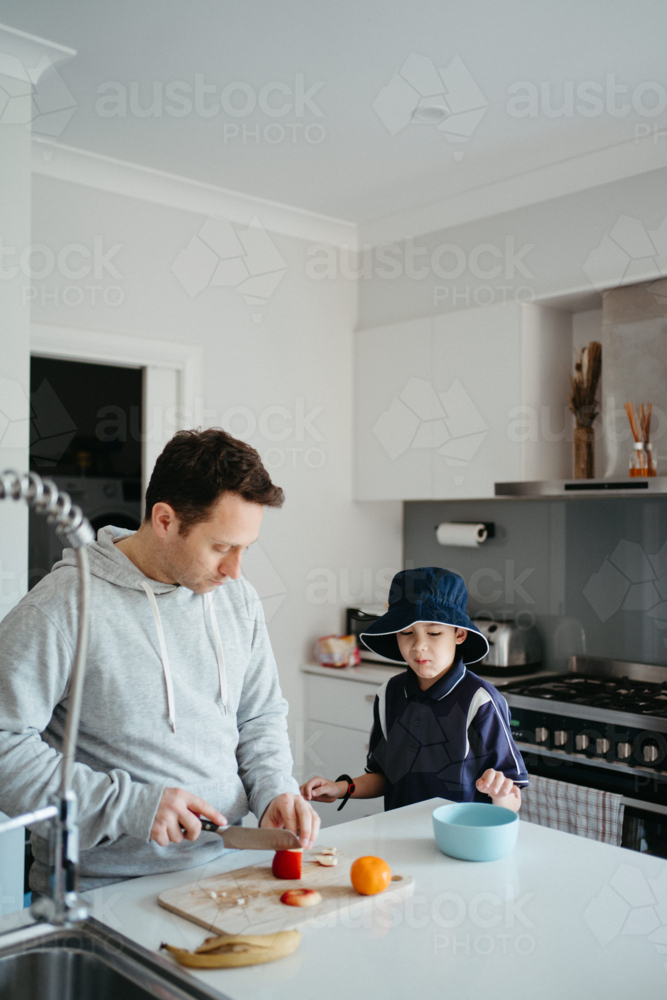 Middle-aged guy cutting fruit for school snacks on the kitchen counter with young boy - Australian Stock Image