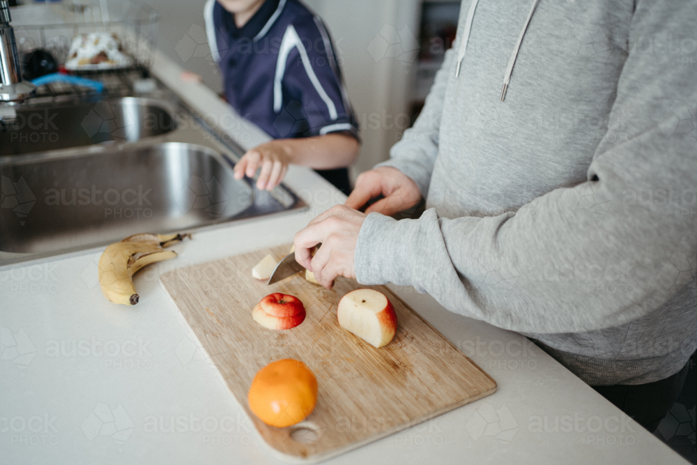 Middle-aged guy cutting fruits on the kitchen counter with young boy waiting beside him - Australian Stock Image