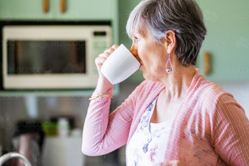 Middle aged woman in kitchen drinking mug of tea - Australian Stock Image