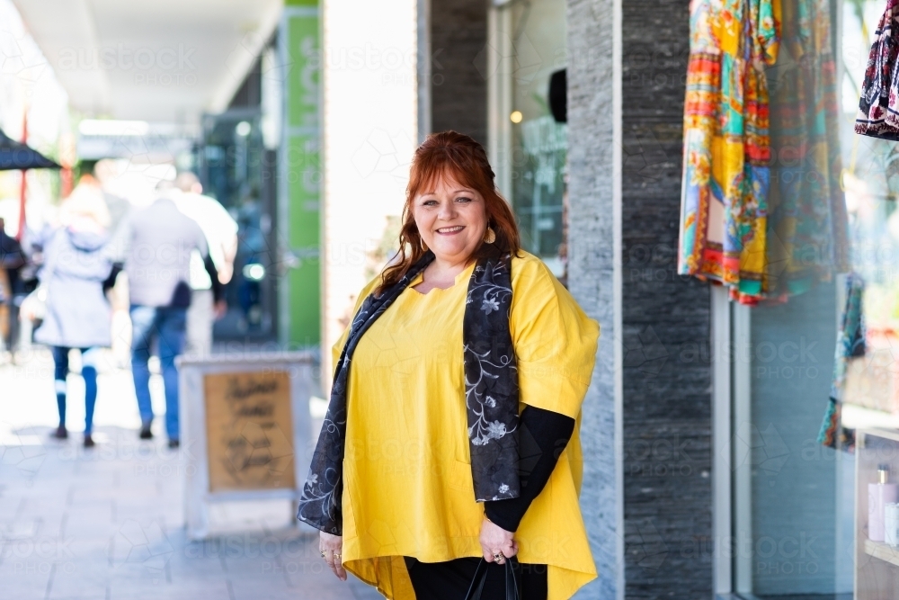 middle aged woman in yellow standing outside of shop smiling - Australian Stock Image