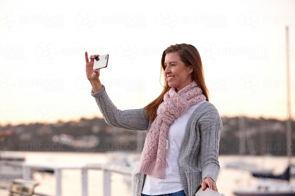Middle aged woman using mobile phone on wharf to take selfie - Australian Stock Image