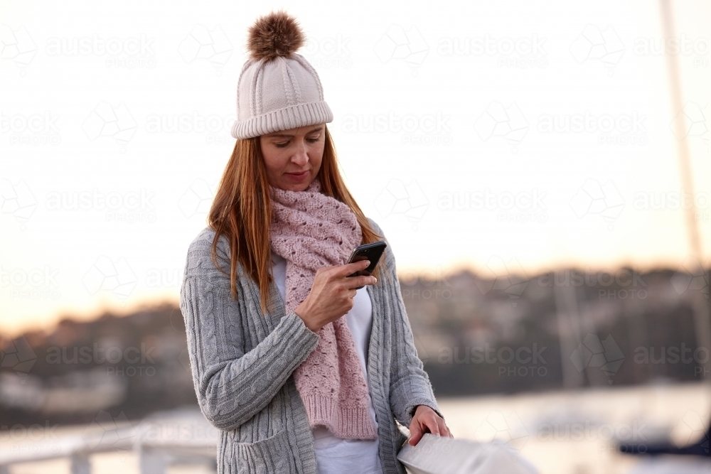 Middle aged woman using mobile phone on wharf wearing beanie - Australian Stock Image