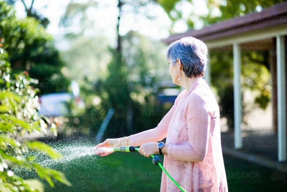 Middle aged woman watering her garden beside her house - Australian Stock Image