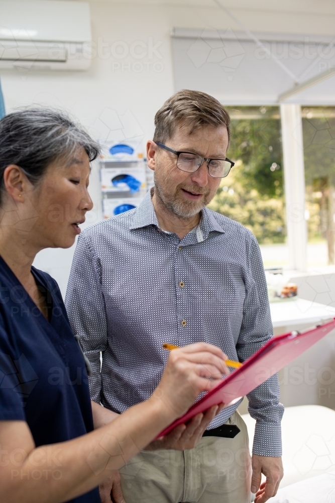 middle aged woman wearing blue scrubs holding a file while talking to a man in a clinic ward - Australian Stock Image