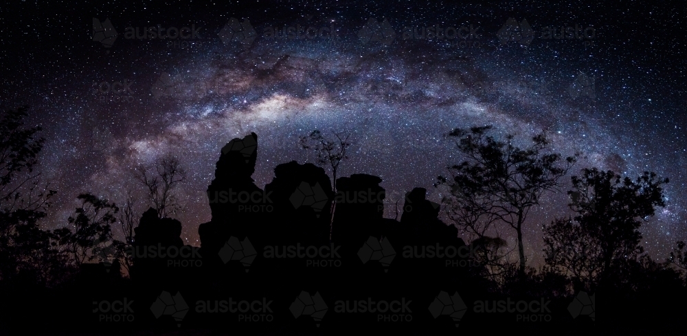 Milky Way galaxy arching over a silhouette of rocky terrain and sparse trees. - Australian Stock Image