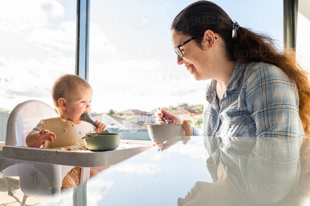 Mother and baby eating breakfast together in morning light with view out window over rooftops - Australian Stock Image