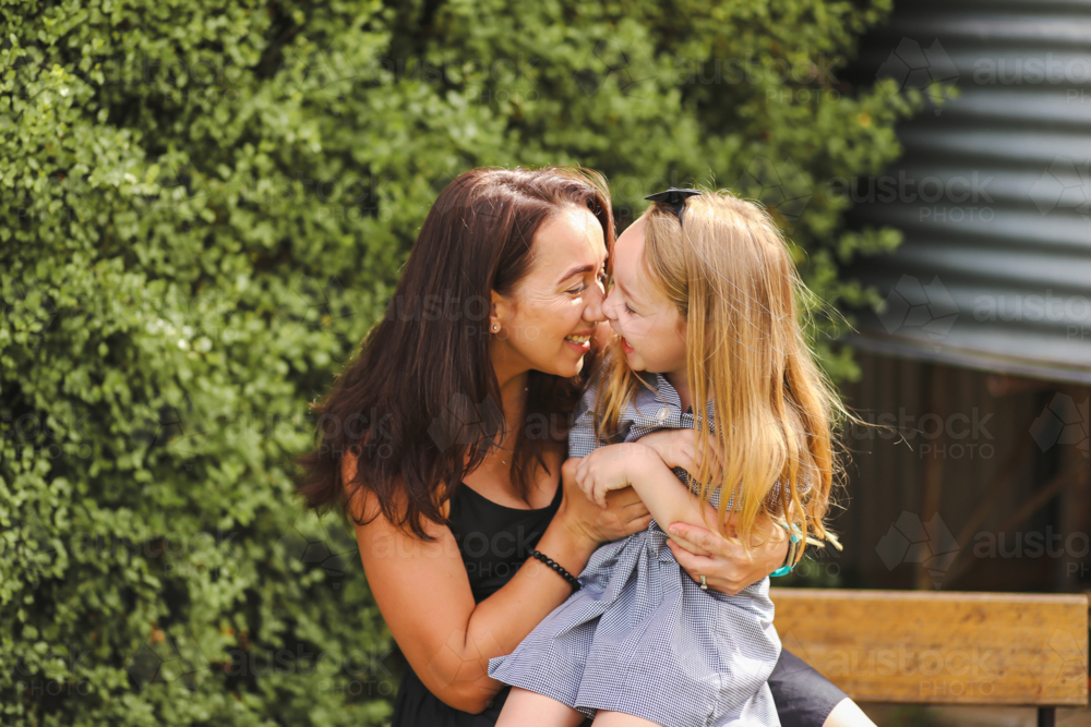 Mother and daughter snuggling in front of green hedge - Australian Stock Image