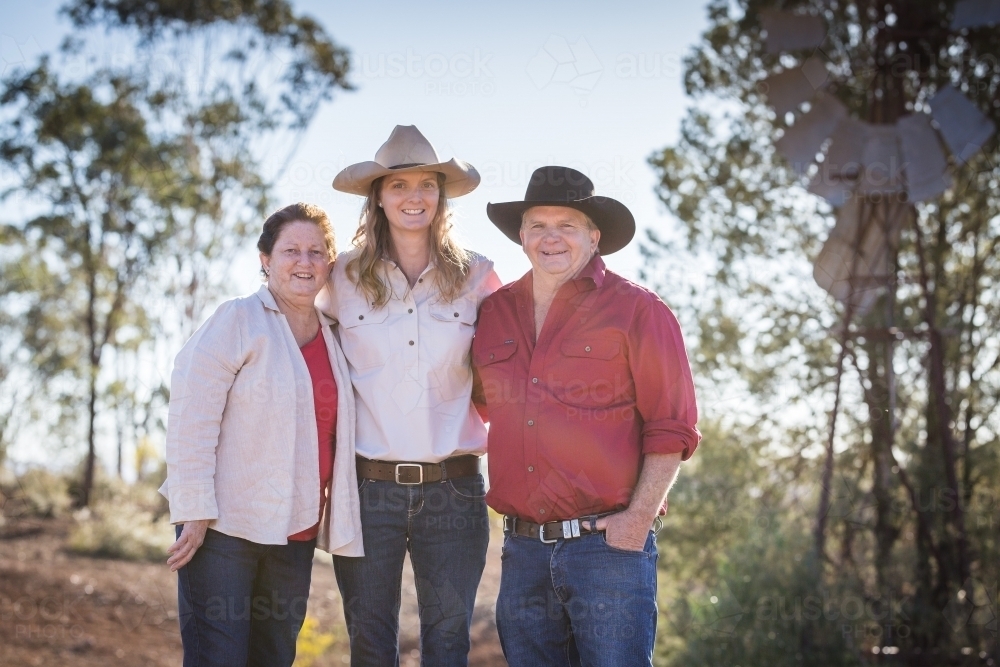 Mother and father standing with arms around daughter with windmill in background on farm in drought - Australian Stock Image