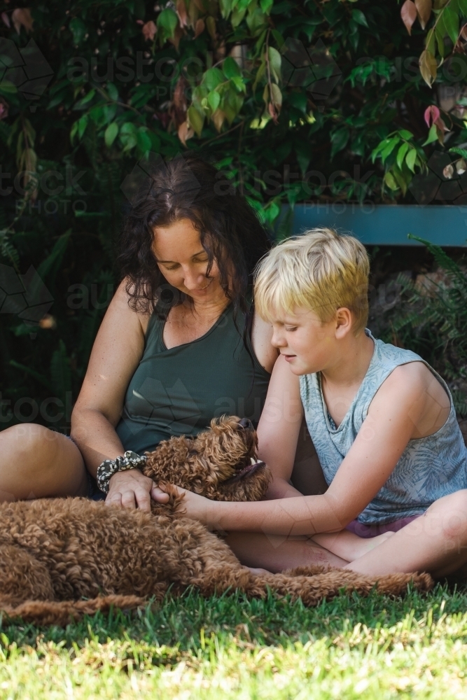 Mother and son sitting on grass with Groodle breed dog - Australian Stock Image