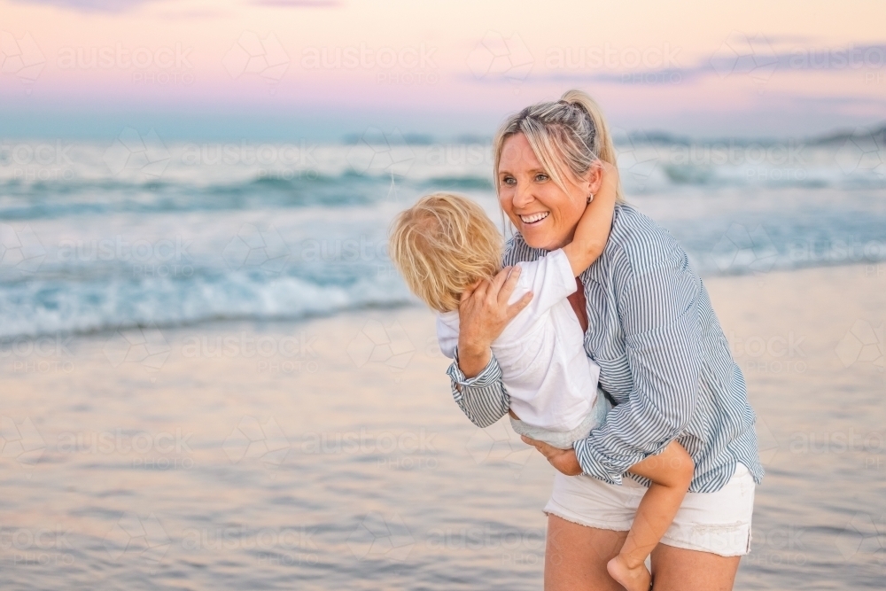 Mother carrying son on the beach in pink twilight light. Sunset on the Gold Coast. - Australian Stock Image