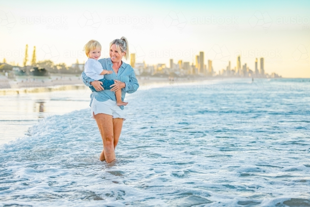 Mother carrying son on the beach with Gold Coast city skyline in background - Australian Stock Image