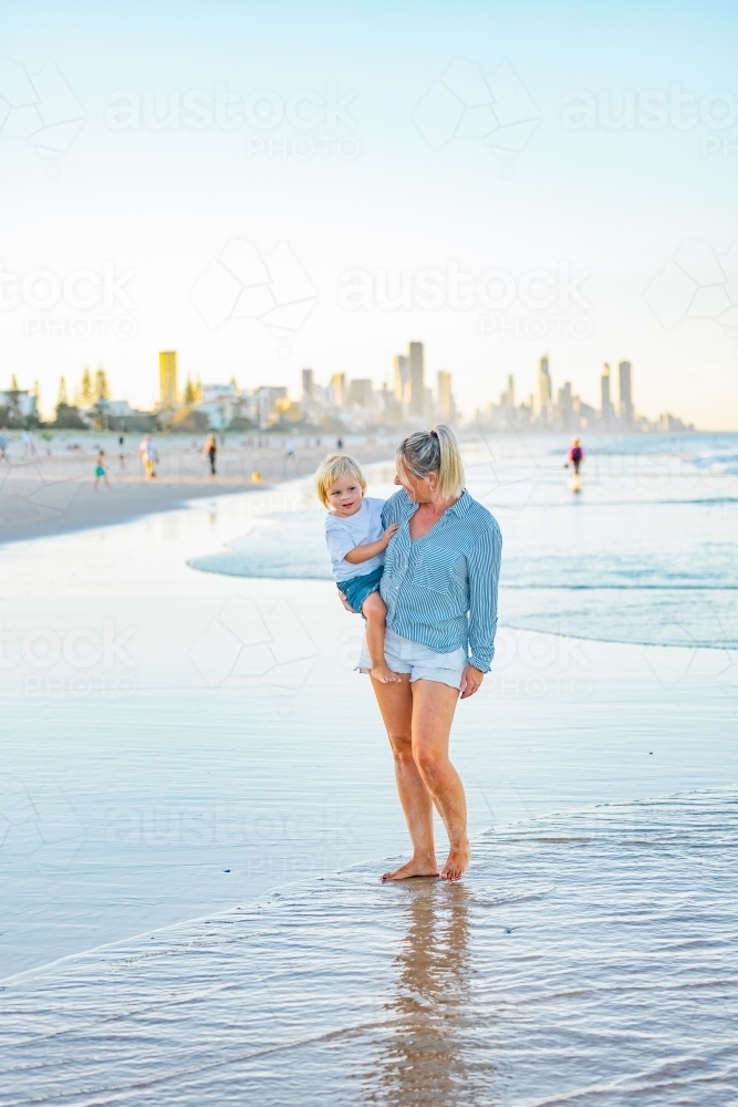 Mother carrying son on the beach with Gold Coast city skyline in background - Australian Stock Image