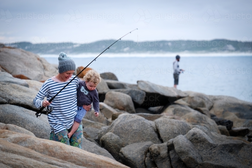 Mother holding child and fishing rod walking over coastal rocks with ocean & fisherman in background - Australian Stock Image