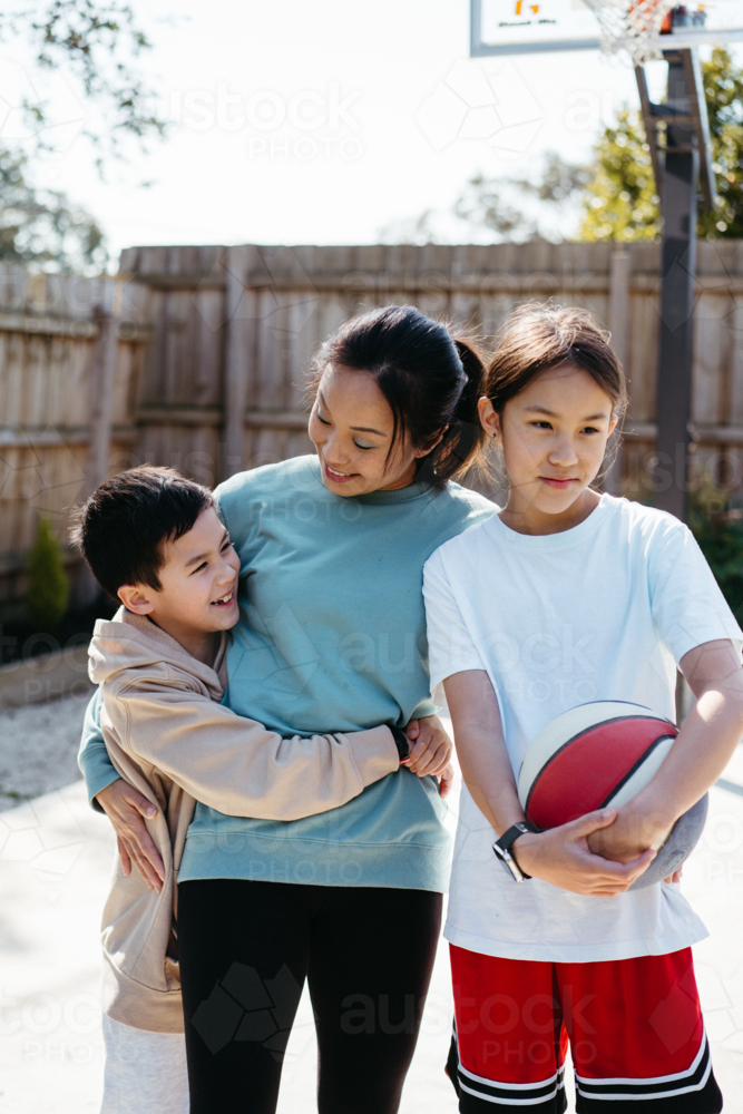 Mother hugs her two young children outside in their backyard. - Australian Stock Image