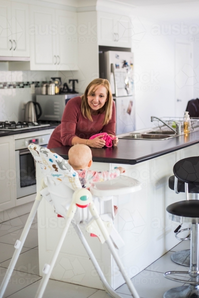 Mother leaning on bench in kitchen smiling at baby in high chair - Australian Stock Image