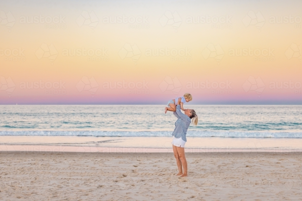 Mother lifting toddler son in the air on the beach with pink sunset sky - Australian Stock Image
