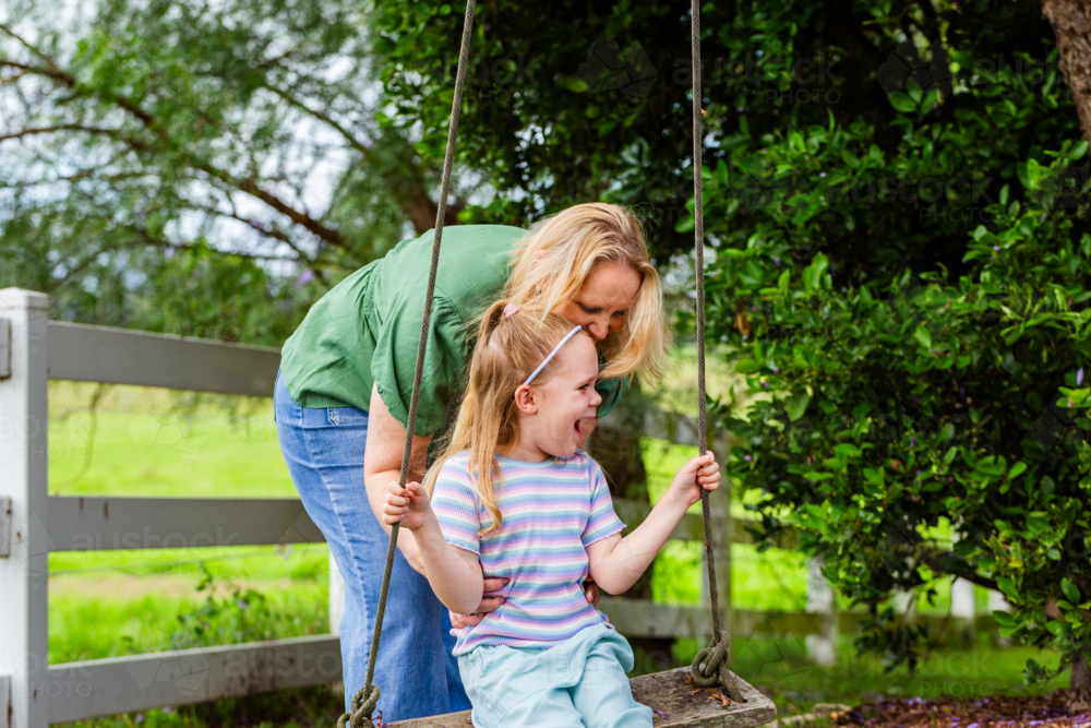 Mother pushing Six year old child on swing in country hanging from jacaranda tree - Australian Stock Image