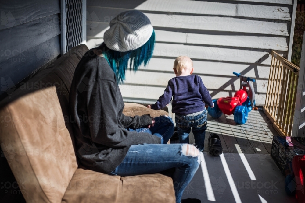 Mother sitting on outdoor lounge watching child play on verandah - Australian Stock Image