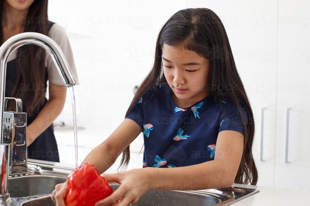 Mother watching over daughter whilst washing vegetable in sink - Australian Stock Image