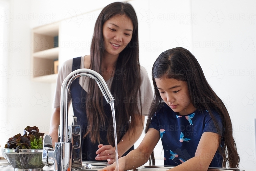 Mother watching over daughter whilst washing vegetable in sink - Australian Stock Image