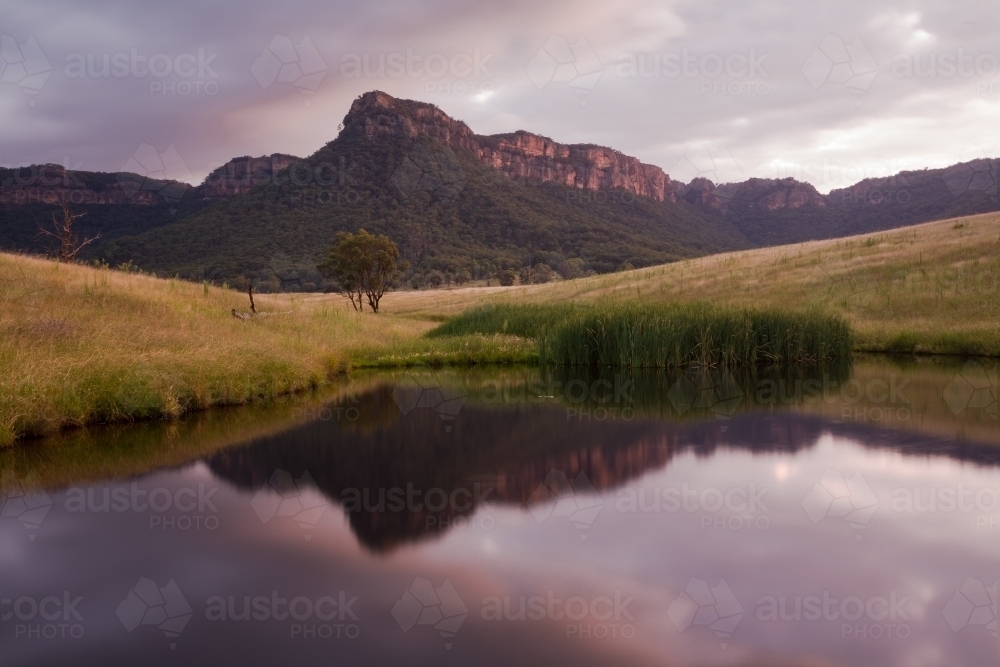 Mountain, paddock and sky reflected in farm dam water at sunset - Australian Stock Image