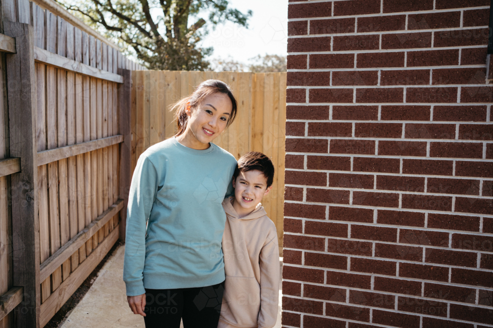 Mum and son standing outside in front of the brick wall of their house. - Australian Stock Image