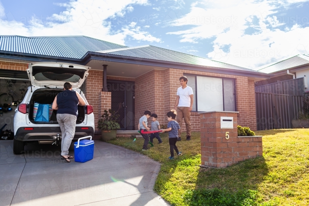 Mum packing family car before sports training with kids playing with hockey sticks on front lawn - Australian Stock Image