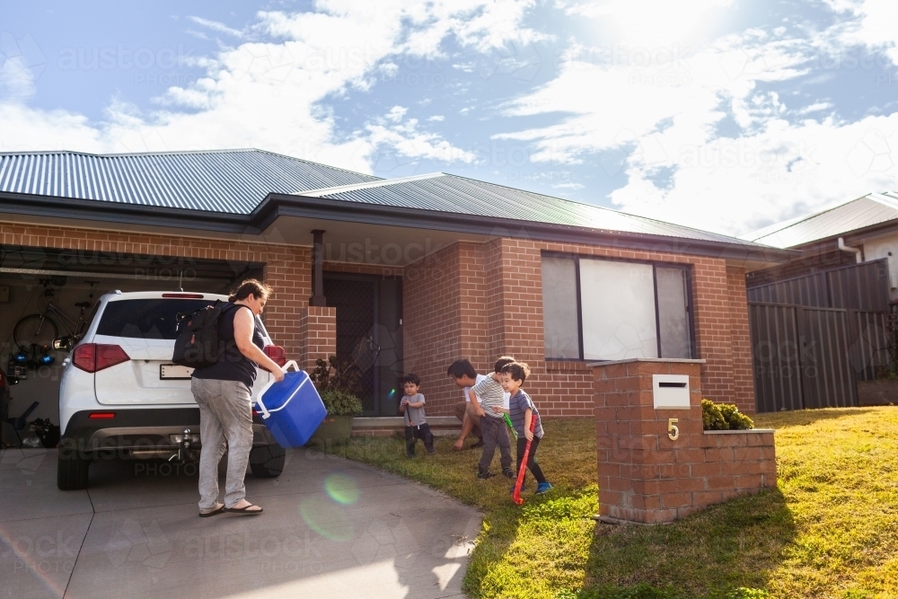 Mum packing family car before sports training with kids playing with hockey sticks on front lawn - Australian Stock Image